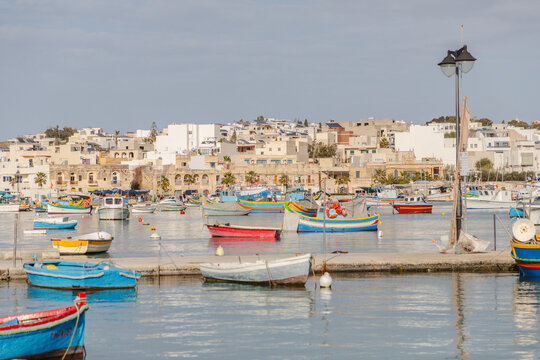 View of vibrant fishing boats bobbing gently on the cerulean water, mirroring the bright buildings under the soft sky, Marsaxlokk, Malta.