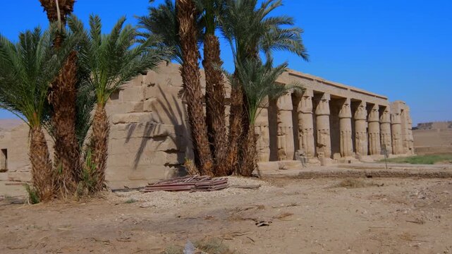 Seti I mortuary temple entrance with palm tree alley and ancient stone ruins in Luxor Egypt. Scenic pathway under vivid blue sky.