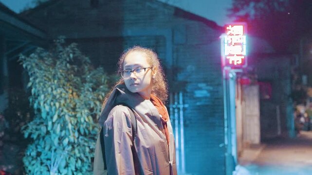 Black woman pausing in neon alley, curly hair backlit and backpack visible, scanning dark passage while distant moped passes cinematic nocturne portrait with bluepink light, tension and quiet