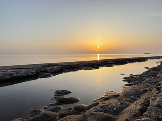 vivid golden sunrise reflects on the glassy calm Baltic Sea, its orange glow lighting up a low-tide shoreline with dark seaweed-covered sand formations and shallow tidal pools in the foreground