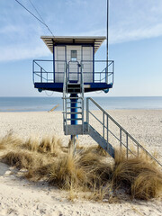 A modern blue and white lifeguard rescue station stands prominently on a wide, sandy beach under a clear blue sky