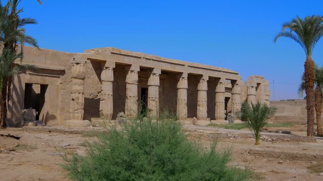 Seti I mortuary temple entrance with palm tree alley and ancient stone ruins in Luxor Egypt. Scenic pathway under vivid blue sky.
