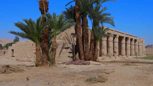 Seti I mortuary temple entrance with palm tree alley and ancient stone ruins in Luxor Egypt. Scenic pathway under vivid blue sky.