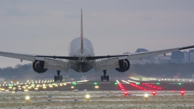 A large passenger jet seen from behind makes a final approach and lands on a lit runway. Tire smoke appears on touchdown as the plane rolls into the distance