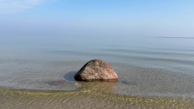 single large dark granite erratic boulder sits half-submerged in a glassy calm sea, perfectly centered against a pale blue hazy horizon, creating a minimalist and meditative coastal composition