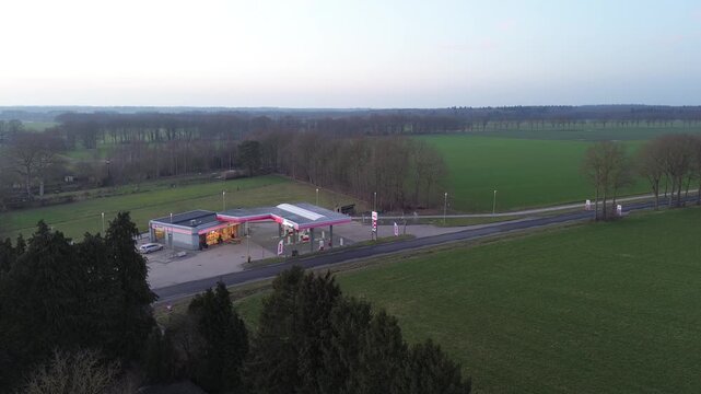 A cinematic drone shot of the N374 provincial road between Westerbork and Schoonloo during dusk. The gas station is illuminated with bright artificial lights, standing out against the darkening