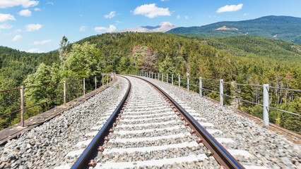 Fototapeta premium Railway track curving through a forested mountain landscape on a bright day, leading towards a rugged peak. Journey and travel concept