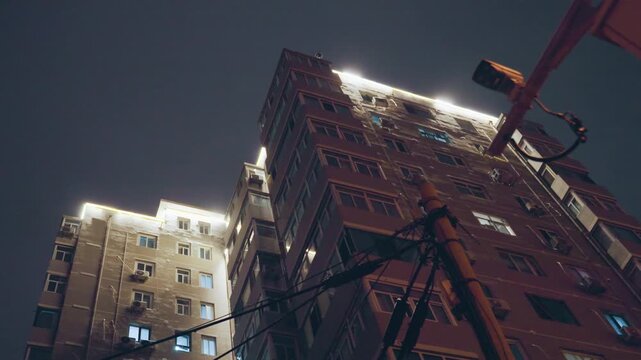 Lit rooftop highrise night sky, string lights along roofline and illuminated windows, power wires and antenna silhouetted against clouded sky, moody film grain and vertical perspective, warm glow