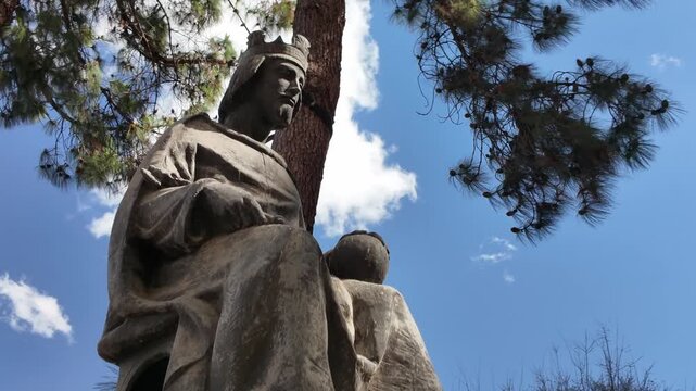 A dynamic low angle view of the historical stone sculpture of King Alfonso X El Sabio framed by green pine trees under a beautiful blue sky with white clouds in Murcia Spain