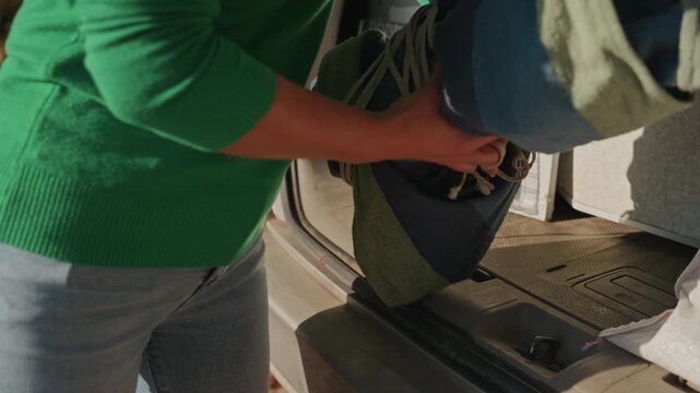 Asian woman loading camping blanket from trunk, rolled blanket tied with rope, green sweater and jeans, open car trunk, autumn leaves on ground, clipboard and gear in back, sunlight warmth, calm