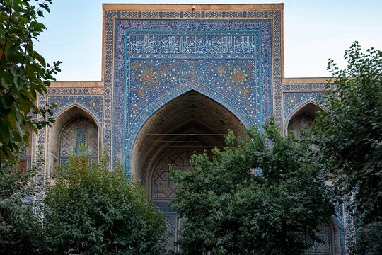 Blue tiled madrasa facade with ornate iwan and garden trees in Uzbekistan