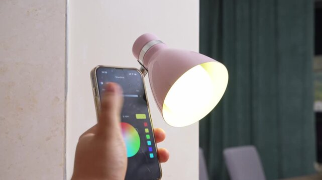 hand holding a smartphone with a colorful smart bulb control app next to a glowing pink desk lamp, modern smart home lifestyle, wireless lighting control, connected device concept, indoor tech comfort