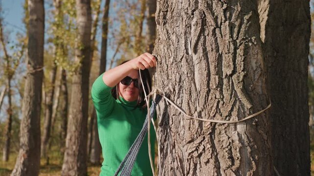 Ensuring hammock stability in forest campsite. Woman ties anchor knot on tree for camping setup. Focused female camper ensures secure attachment of hammock to tree in bright forest