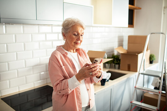 Senior woman using smartphone in kitchen with moving boxes