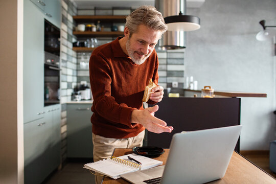 Mature man on video call eating sandwich in home kitchen
