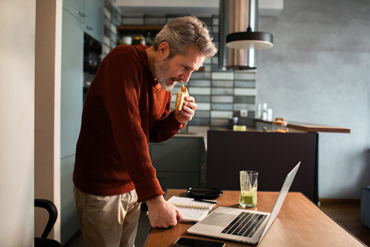 Mature man working on laptop while eating sandwich in home kitchen