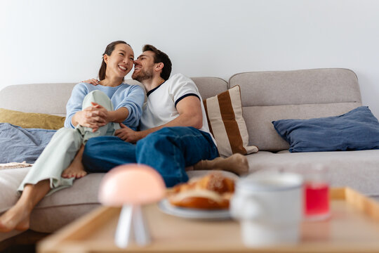 Interracial couple sitting close on a sofa, smiling and leaning in for a kiss. East Asian woman and Caucasian man sharing a tender, romantic moment at home in a cozy, intimate setting