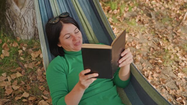 White woman hammock camping reads book, under tree among fallen leaves, green sweater, sunglasses on head, smiling and relaxed, student reader vibe, closeup on pages, warm golden light, peaceful