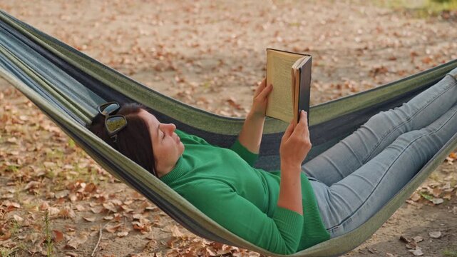 White woman reading in hammock outdoors, green sweater and jeans, sunglasses on head, leafstrewn ground, autumn light, slow living camping break, relaxed leisure vibe, pageturning quiet