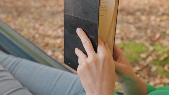 Hammock camping reader holding book outdoors, closeup of hands turning pages while reclining in hammock at campsite, dappled sunlight on leaf litter, denim legs and green shirt sleeve visible, slow