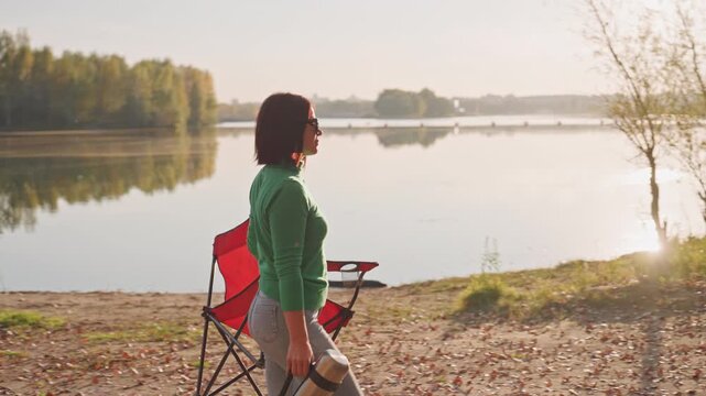 Casual lakeside day with refreshments and relaxation. Woman relaxes beside lake with cozy chair and hot drink. Enjoying peaceful afternoon by water with hot drink and comfy seating