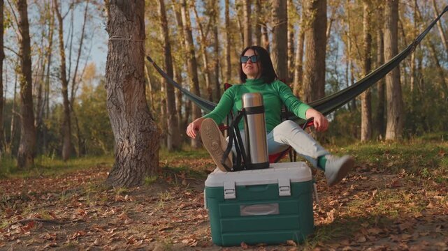 Forest camping woman sipping thermos. Sitting on cooler beside hammock strung between birch trunks, wearing sunglasses, legs propped on cooler, relaxed posture, warm autumn sunlight filtering