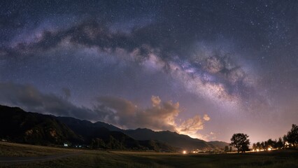 Fototapeta premium A breathtaking night sky showcasing the Milky Way galaxy above a serene mountainous landscape with scattered lights and silhouetted trees.