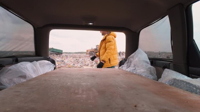 Caucasian Man In Yellow Jacket Lifting Board From Van At Recycling Dump, Straining To Unload Large Sheet, Machinery And Piles Of Waste In Background, Gritty Urban Salvage Scene