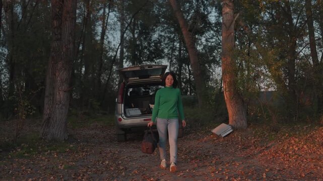 Woman walking from car to lakeside at sunset, green sweater catching warm light, casual stride across leafstrewn path, carrying duffel toward water, pause for reflective gaze over calm surface,