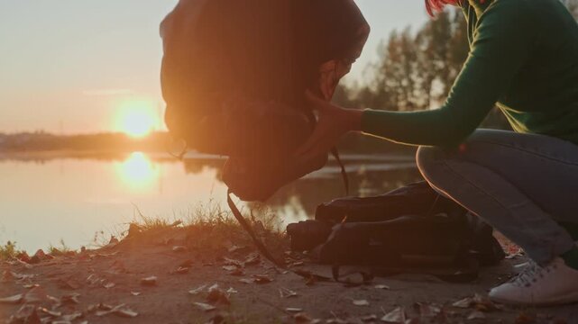 Lakeside sunset camper assembling tent shelter by water, kneeling on shore fitting tarpaulin and groundsheet, securing pegs and ropes, closeup of hands fastening zippers, warm sunset backlight,