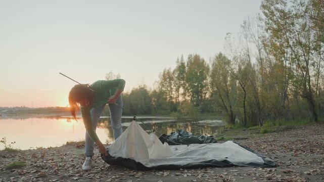 Wide shot white woman pitching tent lakeside sunset, full body silhouette, leafstrewn shore, stretching canvas, securing stakes, soft golden light, calm reflective water, solo camper preparing evening
