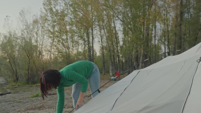 Woman securing tent pegs lakeside at dusk, focused hands hammering stakes into ground, closeup of fabric and poles, methodical campsite prep with tools and rope, instructional practical vibe