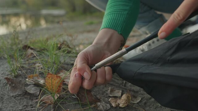 Closeup hands assembling tent pole, white woman securing stake on lakeshore, fabric tension against ground, fingers gripping metal pole, autumn leaves and evening light, focused camping setup