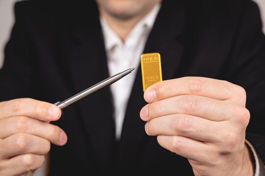 Une main montre un lingot d'or avec un stylo. En arri&egrave;re plan, une personne en costume et chemise blanche.