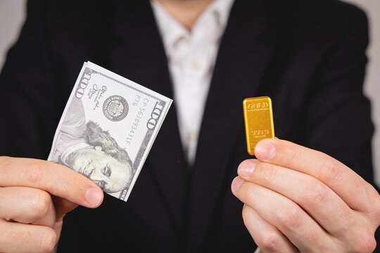 Une personne qui tient un lingot d'or et un billet en dollar dor&eacute;e ses mains. En arri&egrave;re plan, une personne en costume et chemise blanche. Concept de dollar vs or.