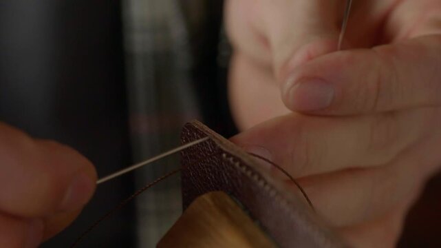 A very detailed close-up shot focusing on the two needles and thick, waxed thread as a craftsman performs a traditional saddle stitch on natural brown leather.