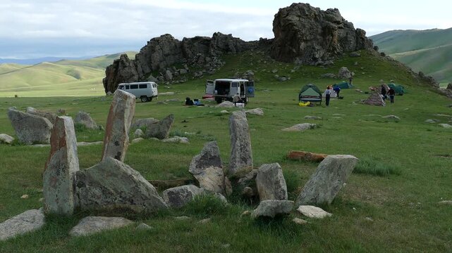  Stone slabs of ancient funerary constructions (Kheregsuurs) and tourist camp
in the Mongolian steppe against the backdrop of the mountains. Summer sunny evening.  Landscape of Mongolia.