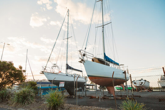 Two sailboats resting on dry land in a boatyard under a cloudy sky