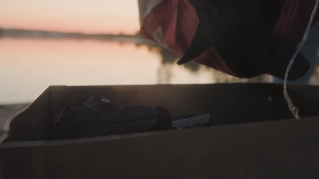 Hands placing charcoal into grill at lakeside sunset, closeup of cardboard box and briquettes, gentle water reflections and warm twilight, fingers arranging fuel for campfire cooking, quiet shore