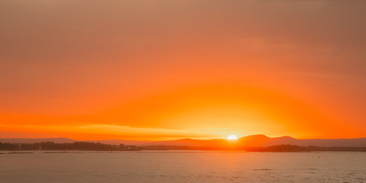 Vibrant orange sunset over a calm body of water with distant hills and a hazy sky