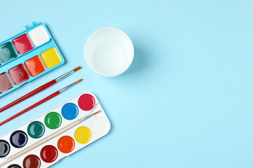 Fototapeta premium Watercolor palette with brushes and glass of water on light blue background, flat lay 