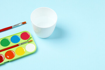 Fototapeta premium Watercolor palette with brushes and glass of water on light blue background, flat lay 