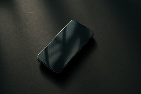 黒デスクに置かれた黒いスマートフォン 画面オフ ミニマルな真上俯瞰 モックアップ背景素材 / Black smartphone with black screen on dark wooden desk, top view, minimal flat lay, mockup background