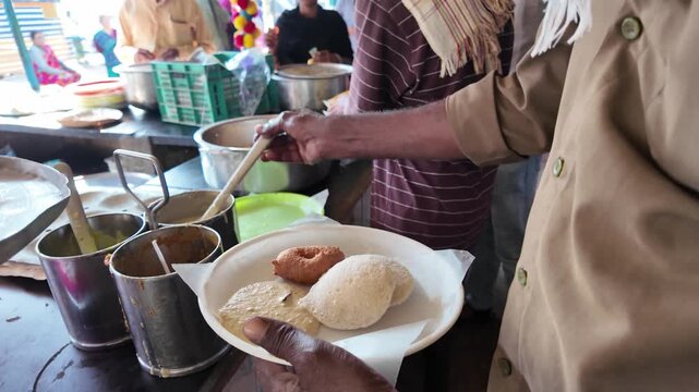 Close-up video frame of a street food vendor serving idli and vada with chutney on a plate at a local Indian food stall. Authentic South Indian breakfast and daily life scene.