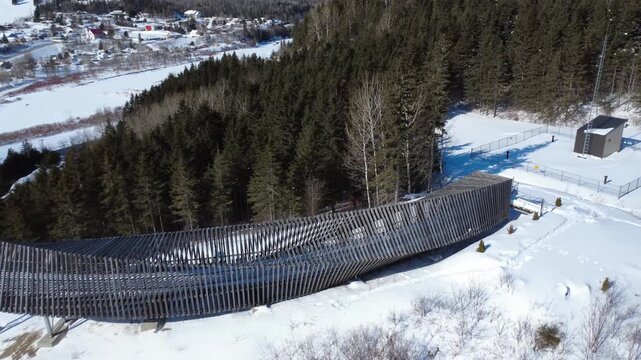 Winter aerial view of a curved wooden footbridge crossing pristine snow next to a dense coniferous forest, showing footprints, a fence, and a small utility shelter. Belvedere des Deux-Rivieres.