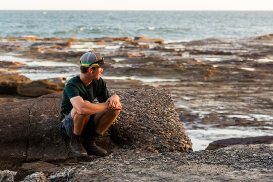 Teen crouched amongst rocks on shoreline looking away in profile with ocean backdrop