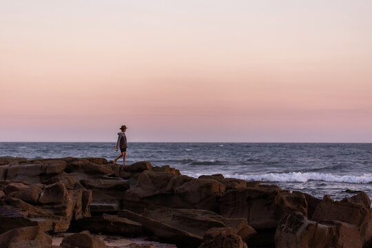 One young person walking alone over coastal rocks in dusk light with flat ocean horizon