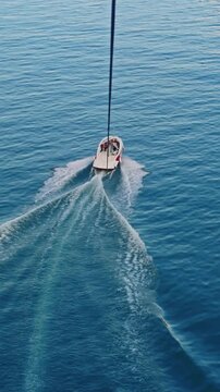 Aerial view of a speedboat pulling a parasail over calm sea, leaving wake trails. Scenic coastline with mountains and city in soft evening light, travel and adventure concept.