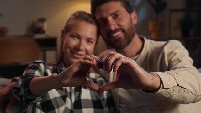 Caucasian middle-aged couple in love showing heart shape with hands enjoying affectionate bonding feelings valentines day cuddling looking at camera happy family husband wife woman man at evening home
