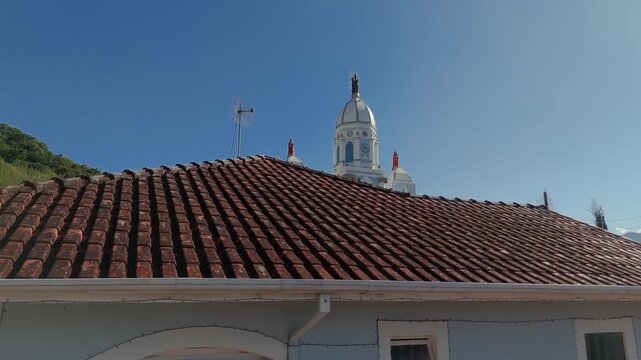 Aerial view of Matriz Church revealed from behind colonial house with red tile roof and green hills in background in Sao Bento do Sapucai, Brazil. Crane up and reveal camera movement